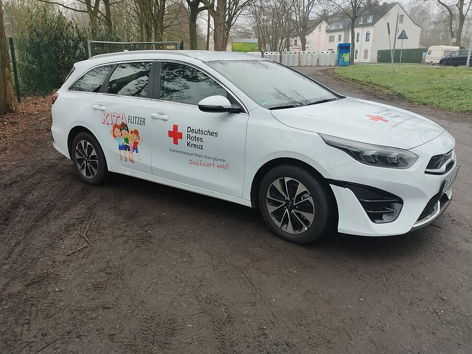 Zu sehen ist ein weißes Auto von der Seite, mit der Aufschrift ,,Kita Flitzer" und dem Logo des DRK Duisburg. Es handelt sich hierbei um den neuen Kita Flitzer