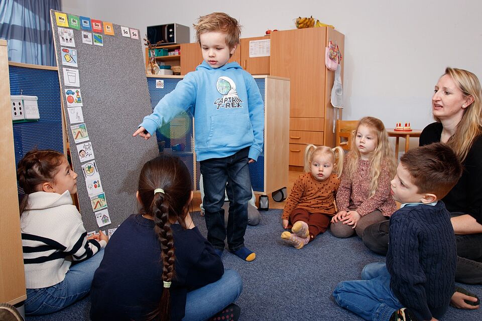 Kinder sitzen im Kreis auf dem Boden zusammen mit einer Erzieherin und besprechen den Tagesablauf 