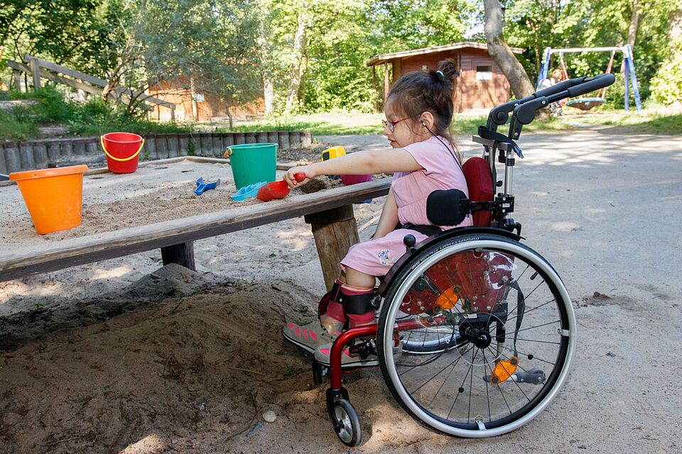 Kind im Rollstuhl sitzt vor einem erhöhten Sandkasten und spielt mit Sand