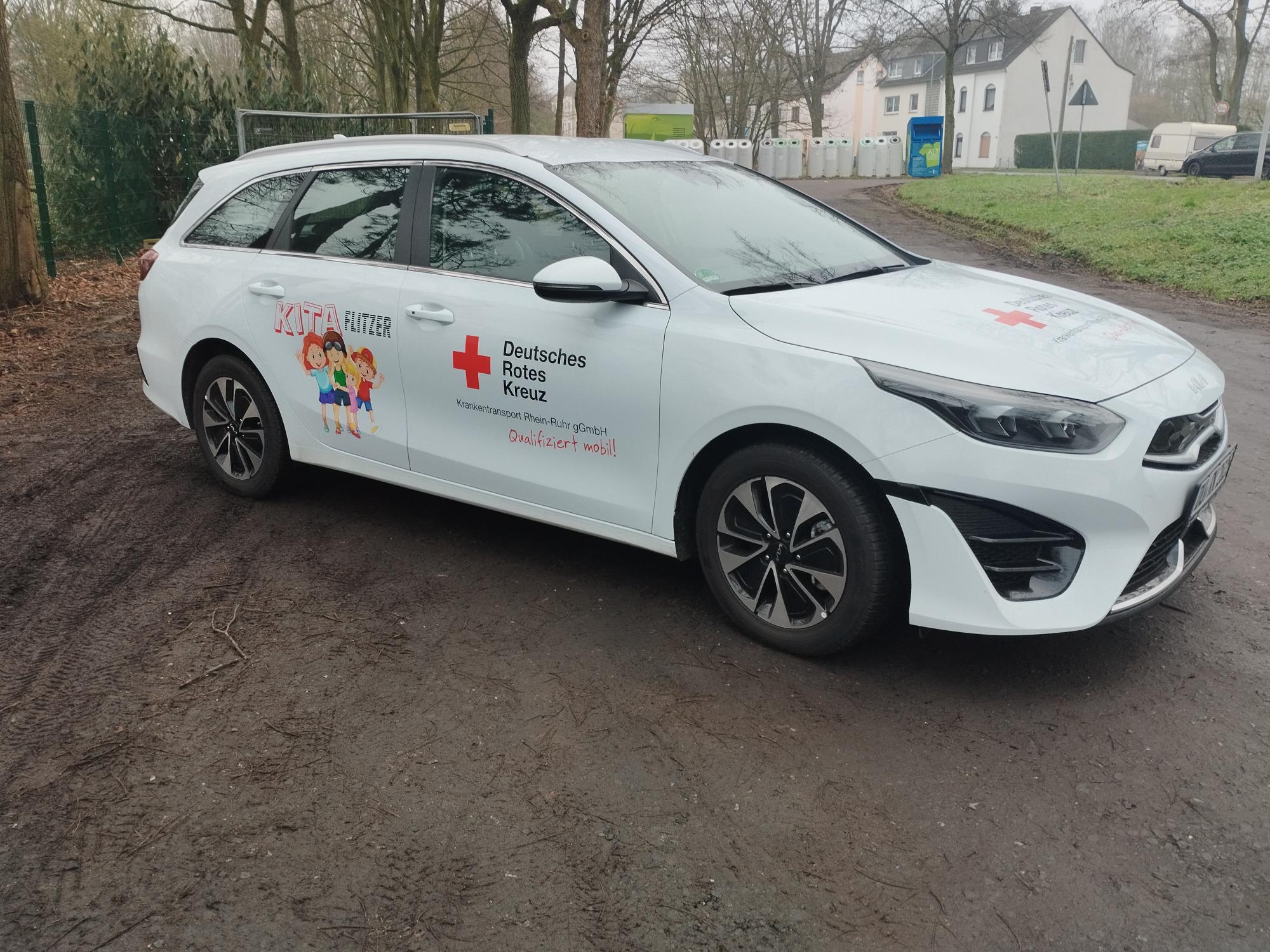 Zu sehen ist ein weißes Auto von der Seite, mit der Aufschrift ,,Kita Flitzer" und dem Logo des DRK Duisburg. Es handelt sich hierbei um den neuen Kita Flitzer